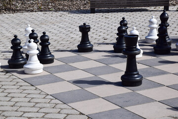  Large outdoor chess set with giant black and white pieces on a stone checkered board in a public park on a sunny day © Olesia