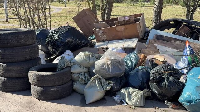 Piled tires and scattered garbage bags on sidewalk, old car tires stacked beside torn bags and cardboard, spring sunlight