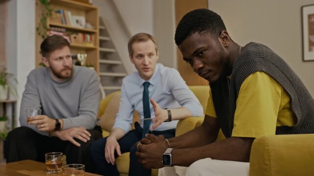 Medium shot of young African American man with clasped hands sitting on chair, nodding and listening to friends supportive speech
