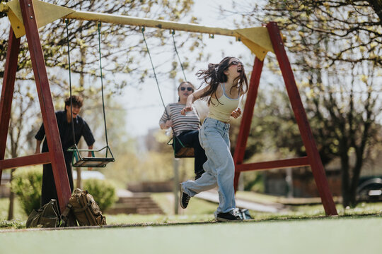 Three friends enjoy playful moments on swings at a sunny neighborhood playground. Young people laugh and run around the swing set during a bright spring day.