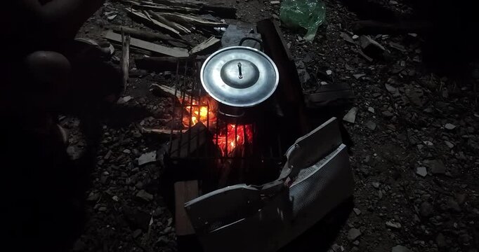 Night shot of a metal pot cooking on a makeshift grill over an open wood fire, surrounded by rubble and scrap debris.