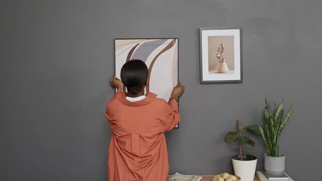 Back view of Black young woman hanging framed abstract art print on dark grey wall above wooden cabinet featuring magazines, bowl of fruit and two potted plants