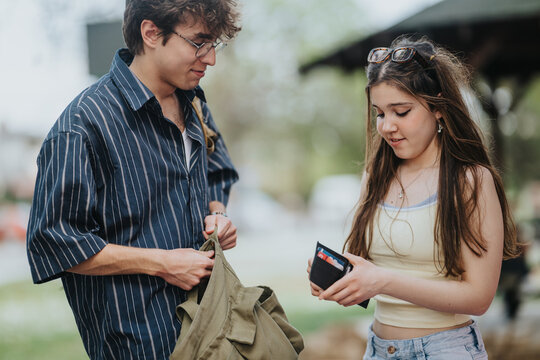 A teenage girl hands money from her wallet to a friend as they stand outside. Two teenagers exchange cash and talk in a relaxed park like environment.
