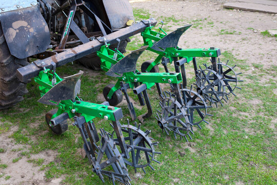 ​A green agricultural cultivator with metal spike wheels is attached to the rear part of a tractor. This mechanical equipment is designed for soil tillage, hilling, and loosening rows in the field.