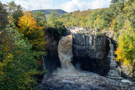 Powerful High Force waterfall plunging through rocky gorge amid autumn trees and misty spray on the River Tees. Scenic landscape and hiking destination. Middleton-in-Teesdale, County Durham, UK.