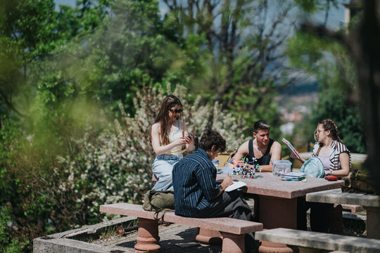 Four students study and chat at a stone picnic table in a green outdoor setting. Young friends review notes and use devices together during a sunny study session.
