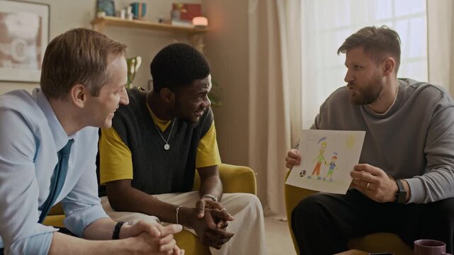 Medium shot of biracial friends gathering in living room to hangout. Bearded man showing sons drawing to friends