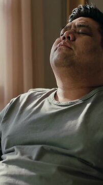 Vertical video: Light lulling man closing eyes, leaning back in chair by window, in tee and pants