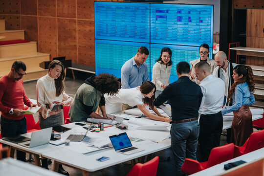 A diverse business team gathers around a table to review plans and spreadsheets. Colleagues collaborate closely in a busy office meeting, brainstorming strategy and next steps.