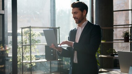 Fototapeta premium Confident Caucasian businessman typing laptop smiling in office morning sunlight professional male leader business man guy entrepreneur manager working online executive boss looking at camera indoors