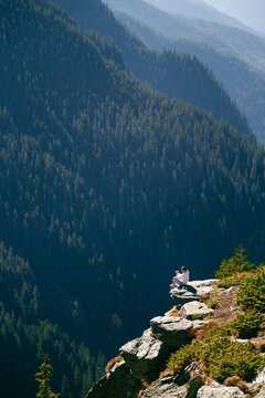 Young woman sitting on cliff edge above deep forest valley in Fagaras Mountains, Romania. Transfagarasan Highway. Freedom, solo travel, mindfulness concept