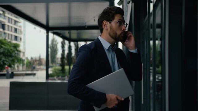 Busy Caucasian businessman male holding laptop talking mobile phone outdoors entrepreneur late business meeting man hurry developer employer guy call smartphone walk city downtown go office building