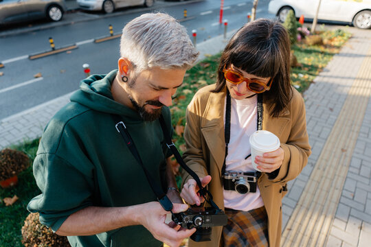 Young couple of photographers checking camera photos while holding coffee on an urban sidewalk