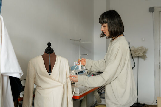 Woman fashion designer measuring and adjusting a cream wrap dress on a mannequin in sewing studio