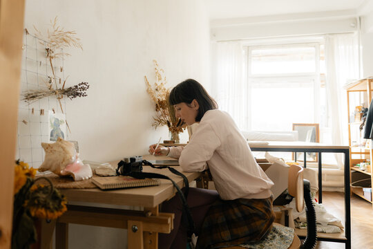 Young woman drawing at a sunlit home studio desk surrounded by art supplies and dried flowers