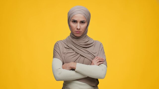 A woman wearing a headscarf stands in front of a bright yellow background. She has a confused look on her face. The scene is clear and captures her expression well in 4k resolution.