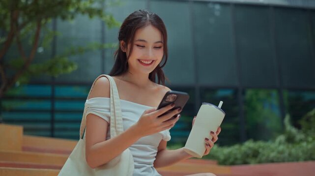Asian woman relaxing under modern eco-friendly building with greenery, smiling while using phone and drinking from reusable bottle, carrying fabric tote bag &ndash; sustainable lifestyle and go green city.