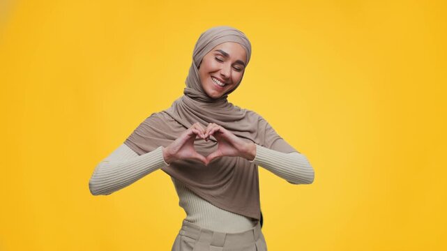 A young woman wearing a headscarf stands in front of a yellow background. She forms a heart shape with her hands and smiles at the camera in 4k video footage.