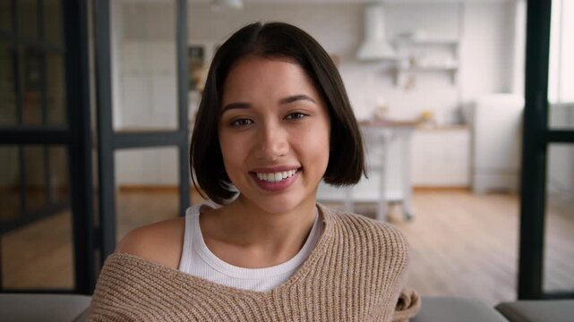 Happy Hispanic woman sitting sofa at home smiling at camera relaxing weekend portrait Caucasian girl female homeowner fall on couch rest toothy smile wife lady housewife tenant own apartment comfort