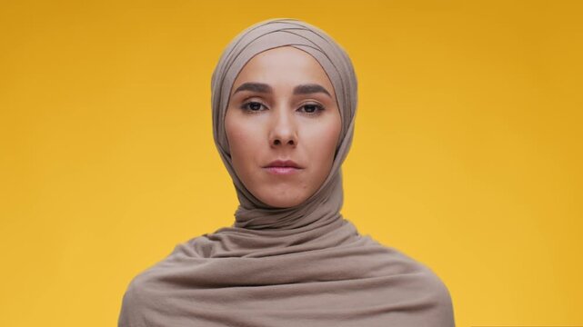 A woman with a hijab stands against a bright yellow background. She has a serious expression while being filmed in high-definition video. The setting looks professional with clear lighting.