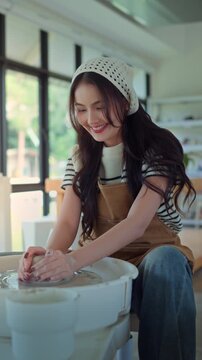 Young asian woman sculpting clay on spinning pottery wheel in ceramic workshop &ndash; relaxing creative hobby, art class activity, and handmade design practice in craft studio environment