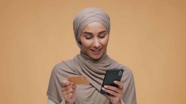 A woman with a hijab holds a smartphone in one hand and a card in the other hand. She smiles while looking at the phone. The background is a simple color. The footage is filmed in 4k.