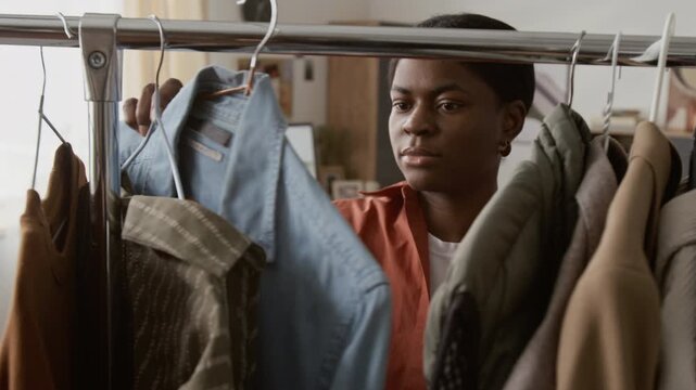 Rack focus view of African American gen Z girl going through clothes on metal rack and choosing denim shirt at home
