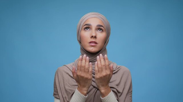 A woman wearing a hijab raises her hands upward in a prayer gesture in front of a blue background. The video captures her expression during the moment.