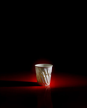 Crumpled white paper cup illuminated by dramatic red lighting against black . Mysterious studio composition creates moody, artistic atmosphere. For conceptual design and creative projects.
