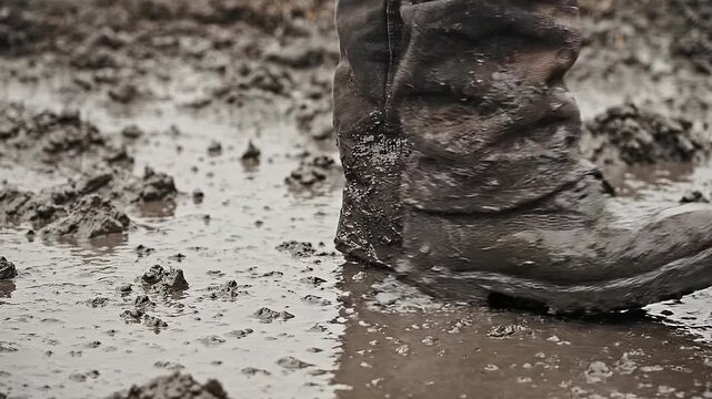 Close up of work boots and pants covered in thick wet mud walking through a puddle. Depicts hard labor, messy conditions, or outdoor adventure. Ideal for construction or agriculture themes.