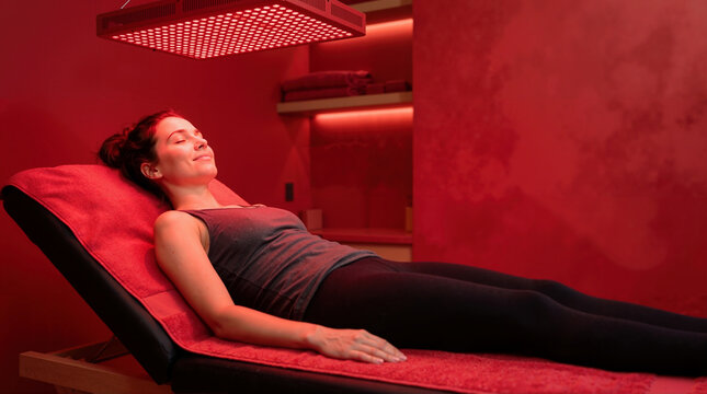 Woman relaxing under a red light therapy panel. Female patient lying on a treatment bed in an immersive red room at a wellness spa