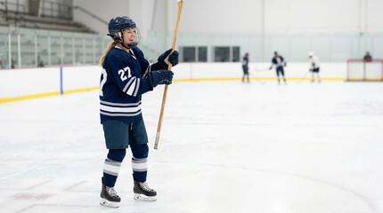 Obraz premium Smiling female ice hockey player standing on rink holding stick. Happy woman athlete in blue uniform and helmet during team practice. Copy space