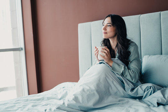 Young woman enjoying a peaceful morning coffee in bed, looking out the window and relaxing in a cozy modern bedroom