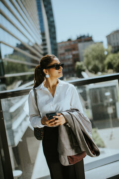 Woman stands outdoors in city while checking phone and holding a jacket