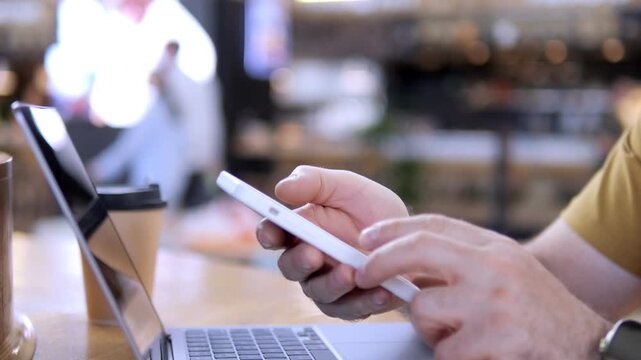 Person uses smartphone and laptop in cafe. Hands hold phone, tap screen. Wristwatch visible. Laptop open beside. Coffee cup nearby. Blurred background with warm lights
