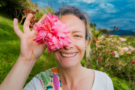 Smiling woman holding pink hibiscus flower over one eye