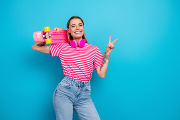 Young woman with pink striped tshirt blue background carries skateboard and wears pink headphones peace sign and smiling in a lifestyle advertising © deagreez