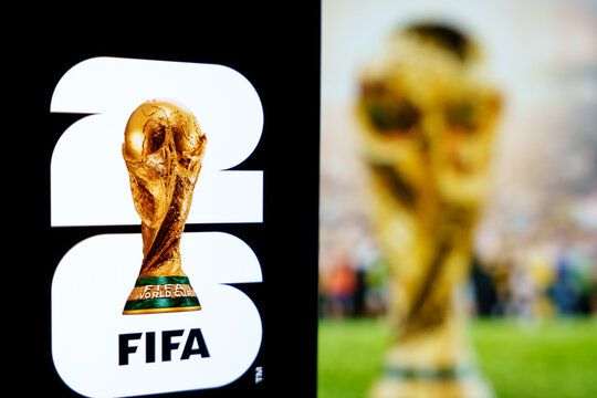 Official FIFA World Cup 2026 brand logo displayed on a digital screen alongside a blurred trophy background, concept of international soccer tournament and football championship merchandise in the USA