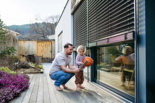 Toddler boy playing with basketball while father controls smart home system on terrace.