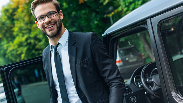 Handsome smiling young businessman coming out of modern luxury car while standing outdoors, confident male entrepreneur in formal suit, corporate lifestyle, success and professional business concept