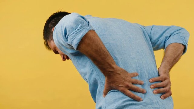 Studio video of a man suffering from severe lower back pain, clutching his lumbar spine in agonizing physical discomfort against a solid bright yellow background.