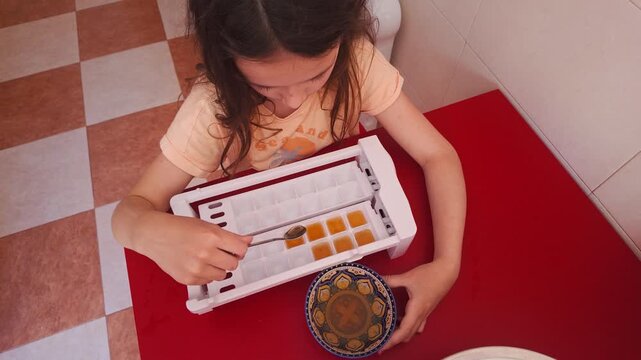 Young girl carefully fills an ice tray with yellow liquid in a home kitchen setting.