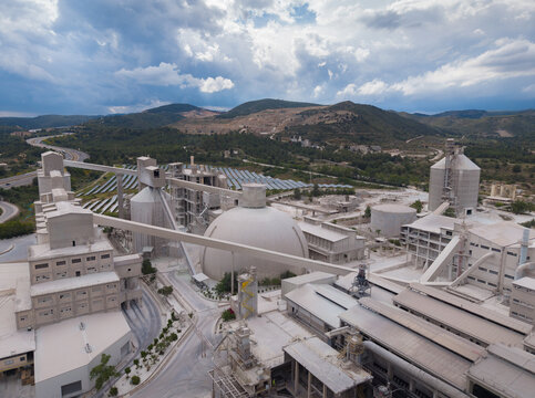 Aerial overview of large cement manufacturing plant featuring prominent spherical clinker dome storage, vertical cylindrical silos, conveyor belts, processing buildings and grinding mills