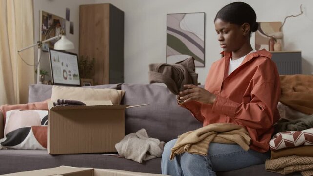 Tilt down view of Black young woman going through garments and placing them into donation box at home