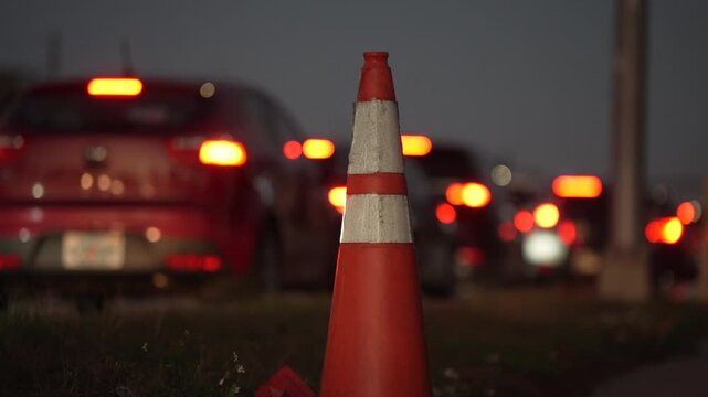 An orange traffic pylon stands on the roadside during evening rush hour with blurred car taillights in the background, showing transport delays