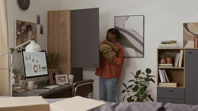Tilt down view of African American girl taking sweaters out of wooden closet and placing them on grey couch next to cardboard box
