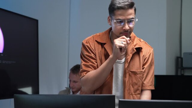 A focused software engineer wearing glasses and gesturing while presenting machine learning workflows and database scripts on multiple office screens.