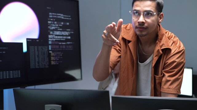 A focused software engineer wearing glasses and gesturing while presenting machine learning workflows and database scripts on multiple office screens.