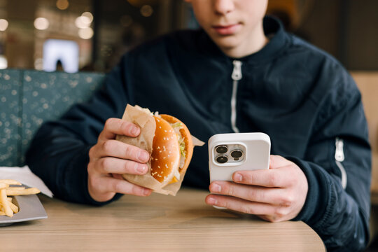 Teenage boy eating burger and using phone in fast food