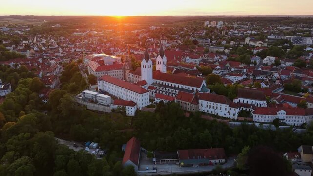 Freising, Germany, viewed from a drone, reveals the Domberg, Freising Cathedral, St. George Church, and old town streets glowing under a summer sunset across the Bavarian landscape.